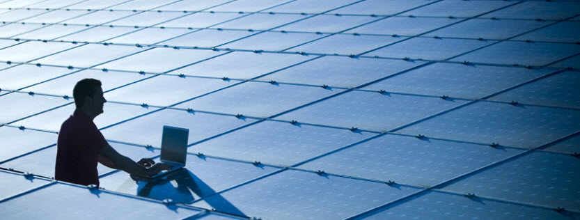 A worker with a laptop is silhouetted against solar panels that are part of a solar farm