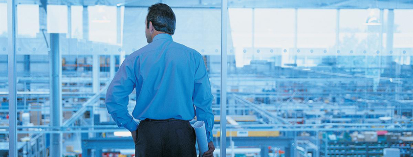 facilities-management Daytime interior view of a man holding a blueprint and surveying the floor of an injustrial facility from an elevated office