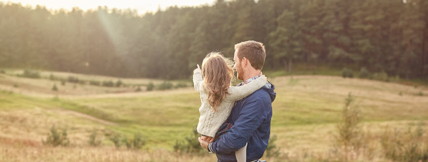 Daytime view of a father holding his young doughter as she points to the sun rising behind trees at the edge of a rolling field