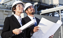 A woman and a man in black suits and white hard hats review blueprints at a construction site