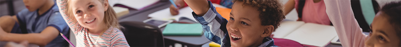 Daytime interior view of young children raising their hands in a classroom.