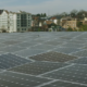 Daytime view of a worker inspecting panels in a solar farm with city buildings in the background