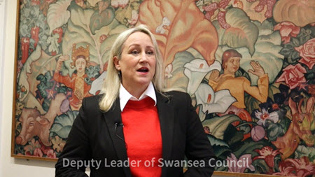 Video screenshot showing Swansea Council Deputy Leader Andrea Lewis in front of a tapestry with the caption Deputy Leader of Swansea Council