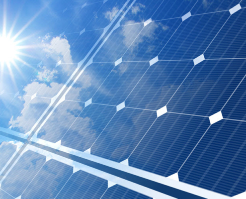 Concept image for solar power shows a closeup of a solar panel superimposed over a sky with clouds and a bright sun